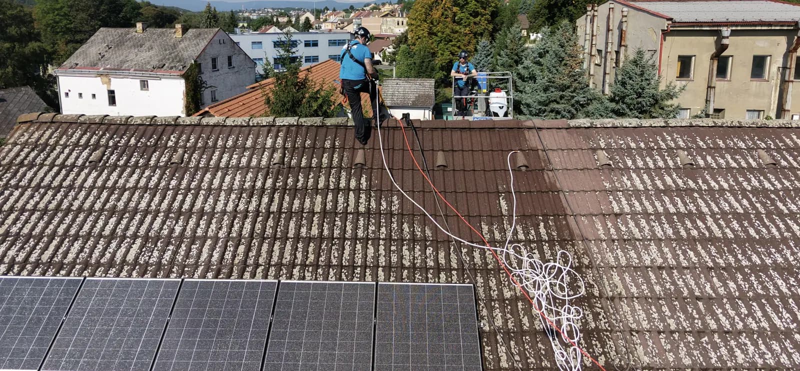 Dva pracovníci s hadicemi čistí střechu z pálených tašek, pohled z dronu